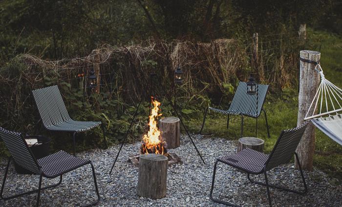 A roaring campfire surrounded by camp chairs set in a rural scene. There is a hammock to one side.